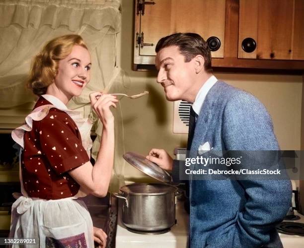 1950s Smiling Housewife At Stove Giving Happy Husband Taste Of Her Cooking.