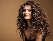 Curly Hair Model. Woman Wavy Long Hairstyle. Brunette Fashion Girl with Volume Hairdo and Natural Make up over Beige Background