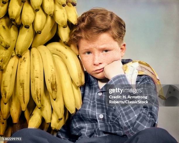 1930s 1940s Distressed Freckle Faced Boy With Stomach Ache Looking At Camera Sitting by Bunch Of Bananas One Half Eaten In Hand.