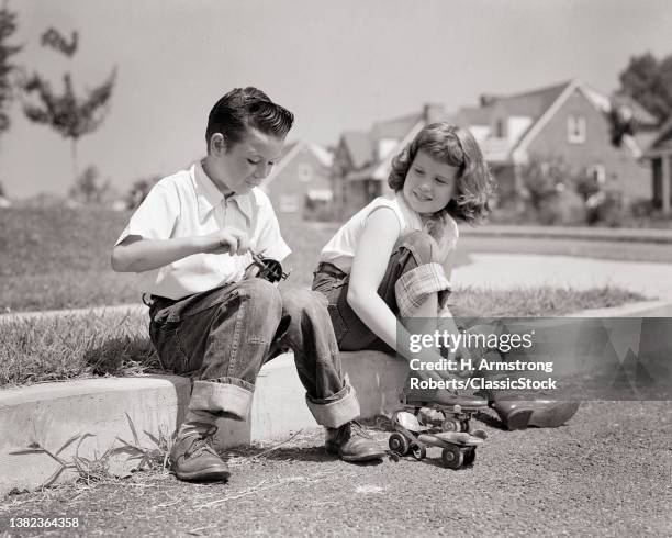 1950s Boy And Girl Brother And Sister Sitting Together On Curb On Suburban Street Putting On Metal Roller Skates.