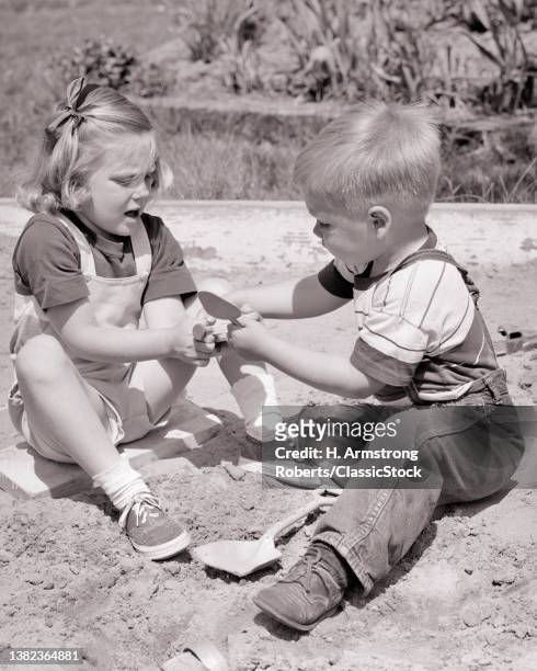 1950s Boy Brother And Girl Sister Sitting Together Playing In Sand Box Arguing Fighting Over Not Sharing Toy Tools Tug Of War.