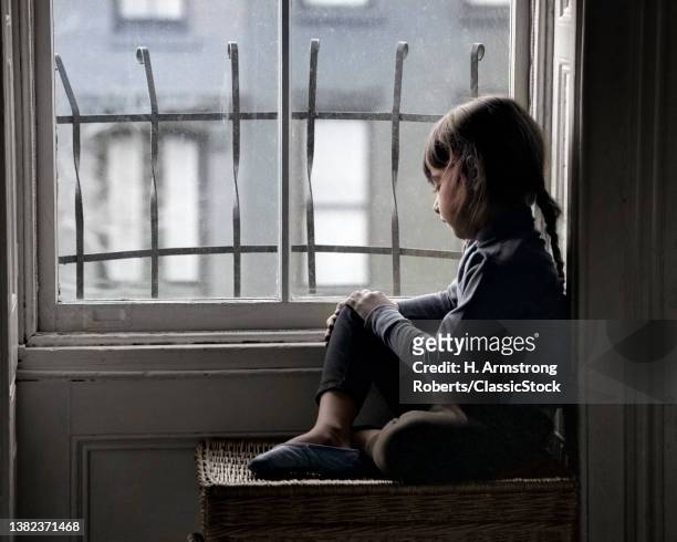 1960s Sad Lonely Little Girl Looking Out Through City Apartment Window With Metal Security Bars.