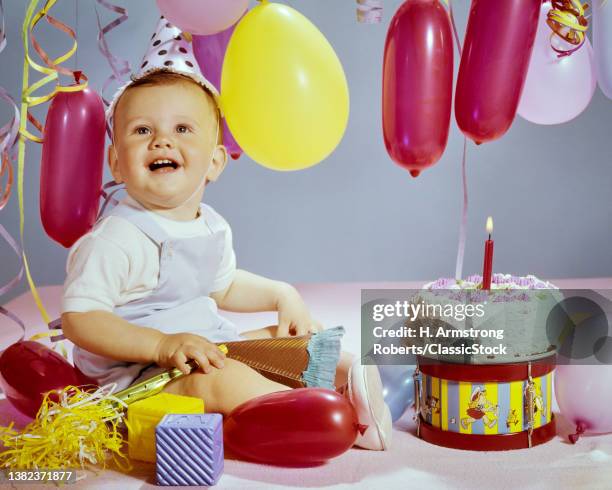 1960s Smiling Laughing Baby Boy Looking Up Sitting Beside Birthday Cake With One Year Candle Red & Yellow Balloons And Streamers.