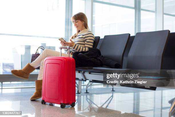 woman waiting at airport departure gate, using mobile phone - equipaje de mano fotografías e imágenes de stock