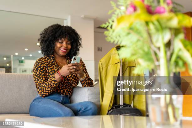 a girl in black ethnic group with hair to the afro with an orange and black shirt. she recently arrived at a hotel to spend the holidays with the travel suitcase. by sending a message with the mobile - project traveller stock pictures, royalty-free photos & images