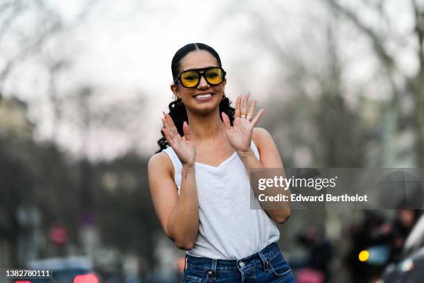 Lena Mahfouf wears yellow sunglasses, gold earrings, a white tank-top, blue denim large jeans pants, silver and gold rings, outside Giambattista...