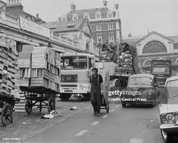 Market worker pulling a sack truck loaded with boxes of produce, with traffic behind him, along a road to the market in Covent Garden, London,...