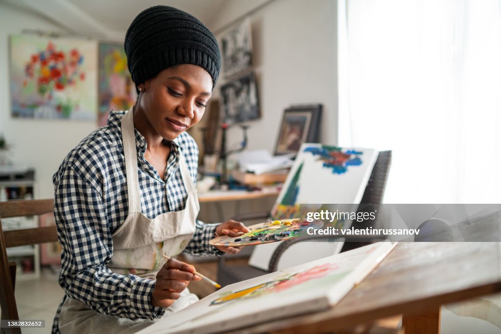 Afro weibliche bildende Künstlerin mit traditionellem afrikanischen Hut
und Schürze, Zeichnen im Kunstatelier