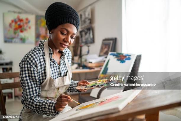 afro female fine artist with traditional african hat 
 and apron, drawing in art studio - schilder kunstenaar stockfoto's en -beelden
