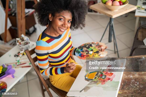 vista superior de la pintura de una artista afro femenina. artista fino sosteniendo una paleta y un pincel, dibujando en un lienzo y mirando a la cámara - art and craft equipment fotografías e imágenes de stock