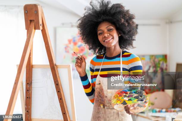 lächelnde afrikanische künstlerin mit afro-haaren und einer schürze, die in einem atelier auf leinwand zeichnet, auf einem stativ platziert - painting artist female stock-fotos und bilder