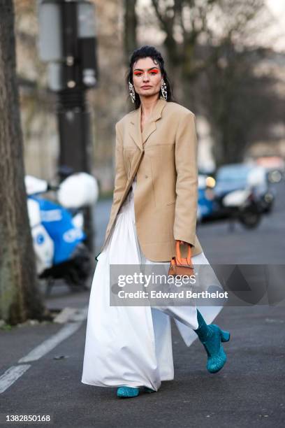 Angela Rozas Saiz wears oversized pearls pendant earrings, a beige long blazer jacket, a white oversized long shirt dress, an orange shiny leather...