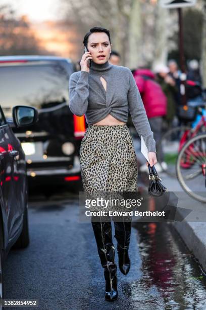 Daisy Ridley wears a gray turtleneck / cut-out chest / knot cropped pullover, a beige with black leopard print pattern knees skirt, a black shiny...