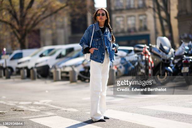 Chloe Harrouche wears black sunglasses, a black sweater, a white t-shirt, a blue denim oversized jacket, a white shiny leather shoulder bag, white...
