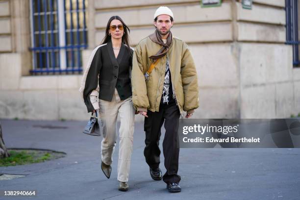 Alice Barbier wears orange sunglasses, gold earrings, a silver large cain necklace, a dark brown and beige oversized buttoned cardigan, a beige...