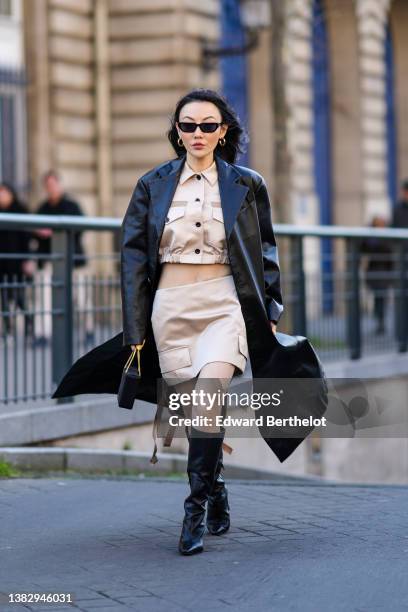 Jessica Wang wears black sunglasses, a black long shiny leather coat, a beige buttoned cropped shirt, a matching beige short skirt, a black shiny...