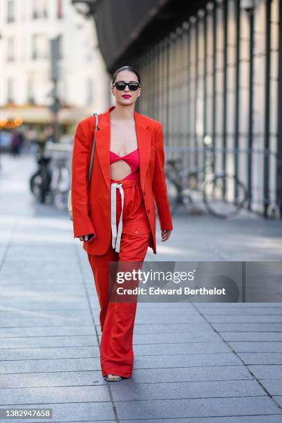 Gabrielle Caunesil wears black sunglasses, a red orange blazer jacket, high waist matching red orange large pants, a red V-neck / cut-out chest body,...