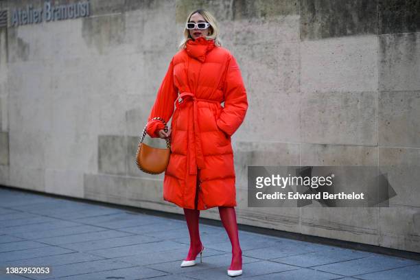Candela Novembre wears white sunglasses, a black turtleneck pullover, an orange oversized long / belted puffer jacket from Stella McCartney, red...