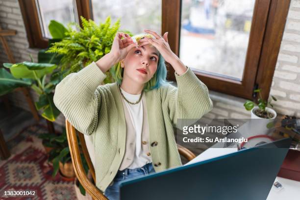 mujer joven con ojo seco usa gotas para los ojos mientras trabaja - bloodshot fotografías e imágenes de stock