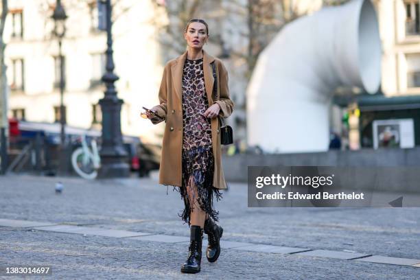 Guest wears a black large hair elastic, gold earrings, a beige with black / brown leopard print pattern sequined / fringed dress, a brown long coat,...