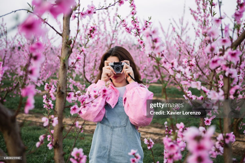 Young woman taking photos of blossoming fruit trees with a retro camera in spring