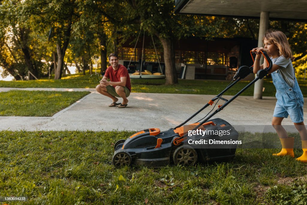 Daddy’s little helper. Boy is mowing the lawn and father supervising him