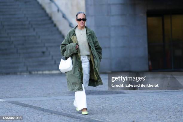Guest wears black sunglasses from Prada, silver chain pendant necklaces, a pale green wool pullover, a dark green oversized long coat, a white shiny...
