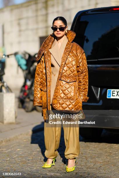 Guest wears black sunglasses, a beige blazer jacket, brown oversized puffy pants, a brown shiny quilted oversized puffer jacket, gold earrings, a...