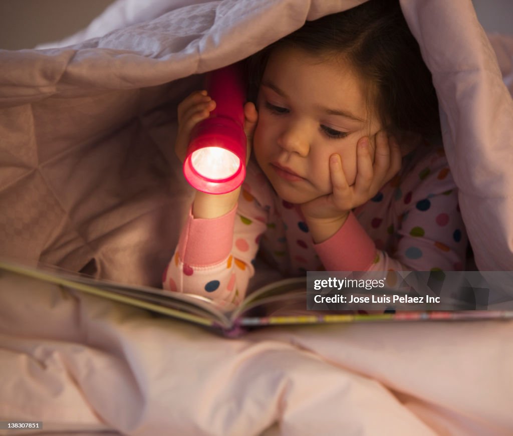 Mixed race girl reading book in bed with flashlight