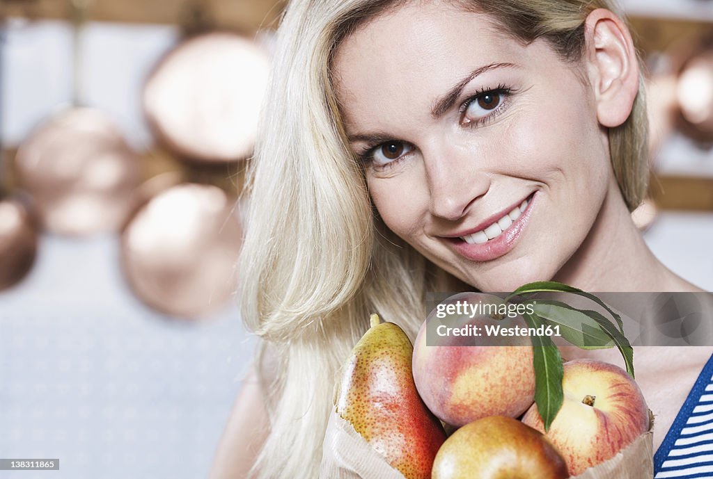Italy, Tuscany, Magliano, Young woman holding fruits in kitchen, smiling, portrait
