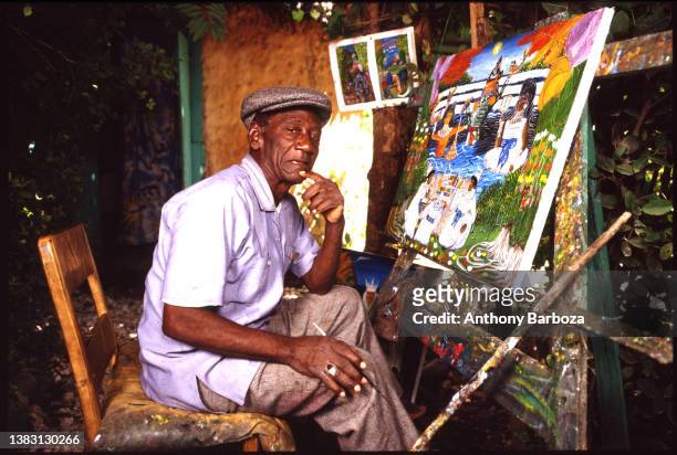 Portrait of Haitian artist Andre Pierre as he sits in front of an easel with one of his paintings, 1991.