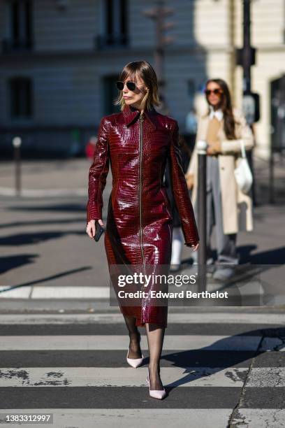 Anya Ziourova wears black sunglasses, a burgundy shiny leather crocodile print pattern long sleeves / zipper midi dress, black tights, pale pink...