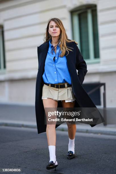 Monica Ainley wears gold and rhinestones pendant earrings, a blue shirt, a navy blue / black long coat, a black shiny leather belt from Miu Miu, a...