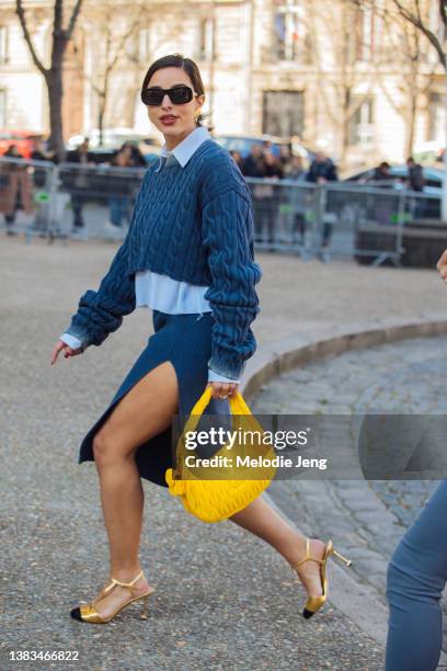 Bettina Looney wears black sunglasses, a blue Miu Miu cropped knit sweater, blue shirt, open-slit blue skirt, yellow Miu Miu bag, and gold heels at...