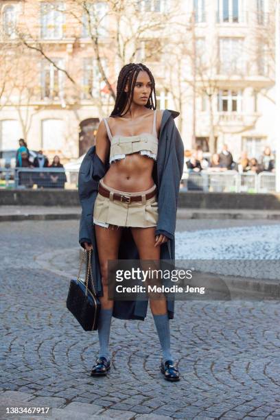 Model Cindy Bruna wears a Miu Miu khaki bra crop top with a matching khaki pleated mini skirt, brown leather belt, gray jacket off her shoulders,...