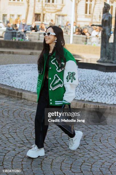 Guest wears a green varsity Louis Vuitton jacket, black pants, black purse, chunky white sneakers, chunky white sunglasses, and hoop silver earrings...