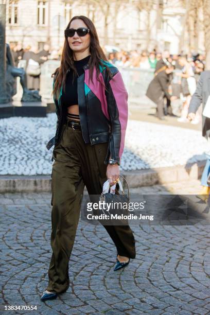 Ece Sukan wears black sunglasses, a black, purple, and green motorcycle leather jacket, black crop top, green pants, black heels, and black and white...