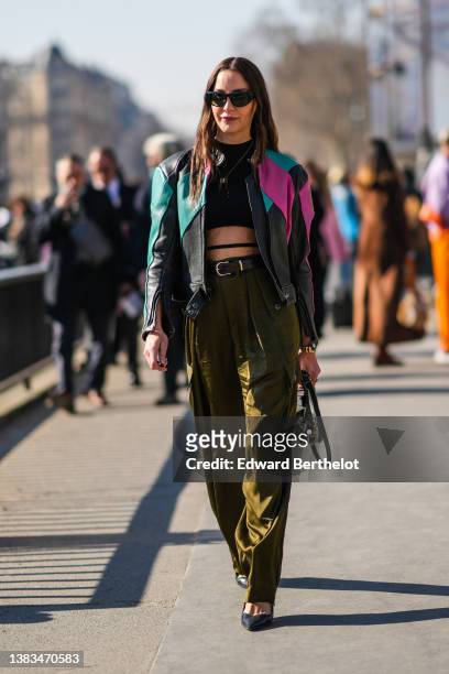 Ece Sukan wears black sunglasses, a black cropped top with laces waist, a black shiny leather with green and pink yoke zipper biker jacket, a black...