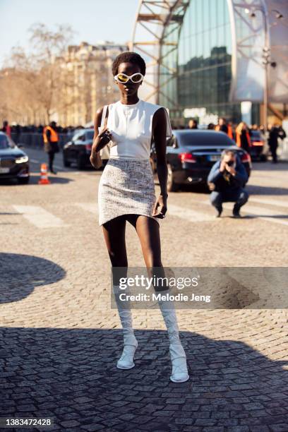 Model Anok Yai wears white alien sunglasses, a white leather assymetrical tank top, white purse, cream print skirt, knee-high white leather lace up...