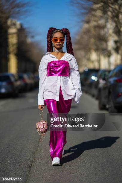 Debbie @blackhey wears a gold flower pattern headband, gold large earrings from Givenchy, orange sunglasses, a white oversized shirt, a neon pink...