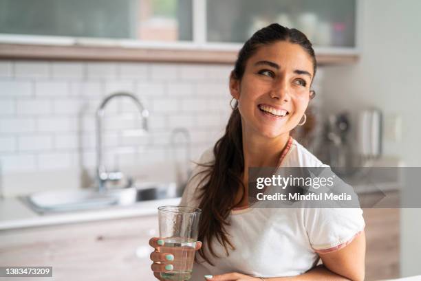 schöne lächelnde junge erwachsene hispanische frau mit einem glas wasser in der küche - drinking water glass woman stock-fotos und bilder