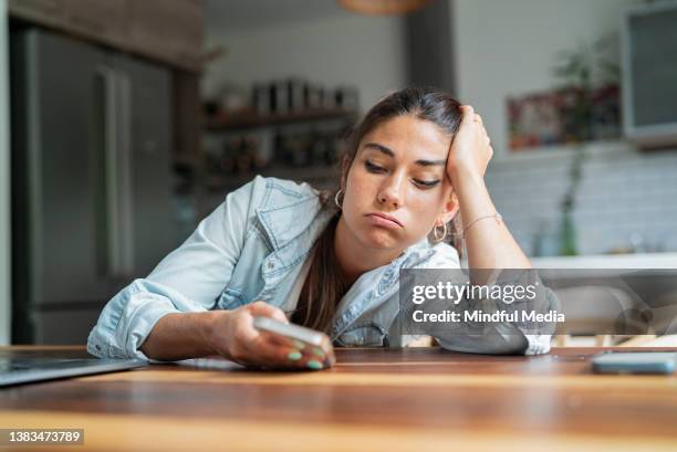 mujer hispana adulta joven aburrida mirando un teléfono inteligente mientras se apoya en la mano - mujer aburrida fotografías e imágenes de stock