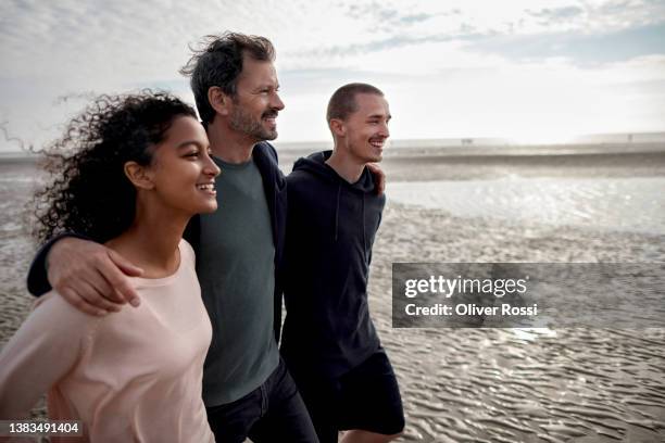 mature man walking with arm around man and woman on the beach - diverse family stock-fotos und bilder