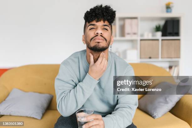 young man with throat pain sitting on sofa at home - throat photos - fotografias e filmes do acervo