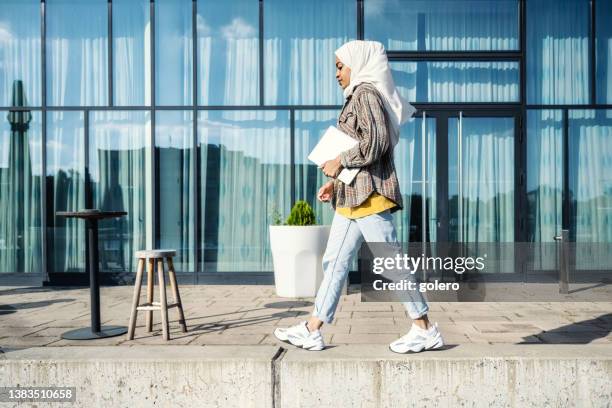 junge frau mit hijab läuft mit laptop vor glasfassade - walking side by side stock-fotos und bilder