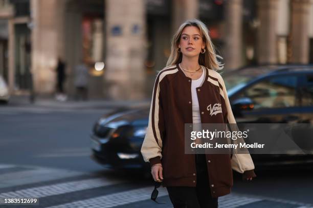 Millane Friesen wearing black shades, a brown college jacket, white top, black pants and golden jewelry on March 07, 2022 in Paris, France.