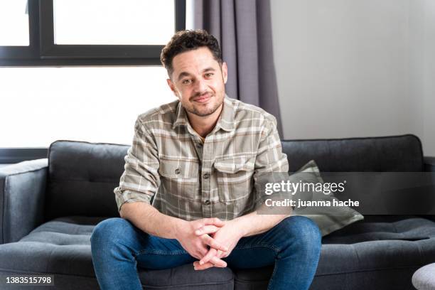 attractive man sitting on sofa at home looking at camera, window and curtains in background. - man sitting on sofa fotografías e imágenes de stock