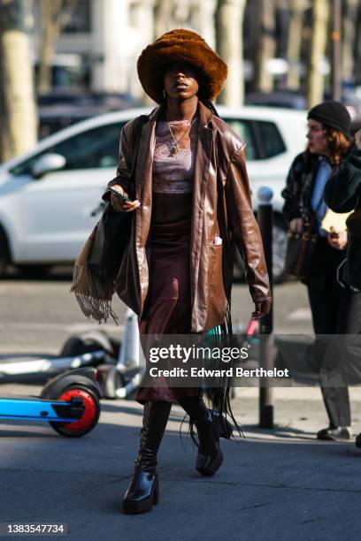 Guest wears a brown oversized fluffy hat, gold earrings, a gold chain pendant necklace, a pink and brown print pattern t-shirt, a brown shiny leather...