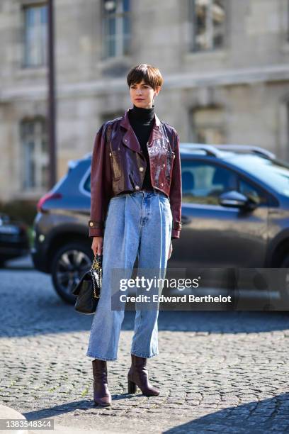 Guest wears a black turtleneck pullover, a burgundy shiny leather cropped biker jacket from Chanel, high waist blue faded denim large pants, a black...