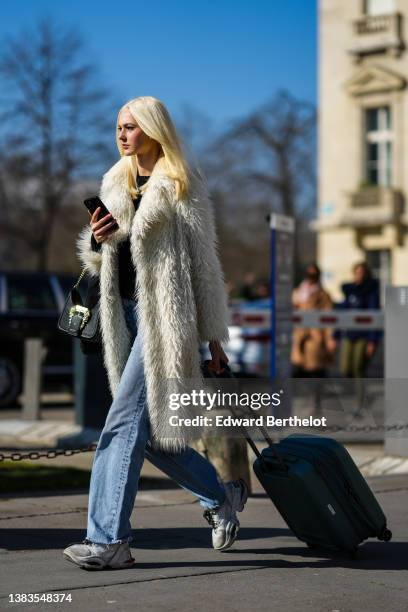 Guest wears a black t-shirt, a white long fluffy coat, blue faded denim wide legs pants, a black shiny leather handbag from Versace, a navy blue...